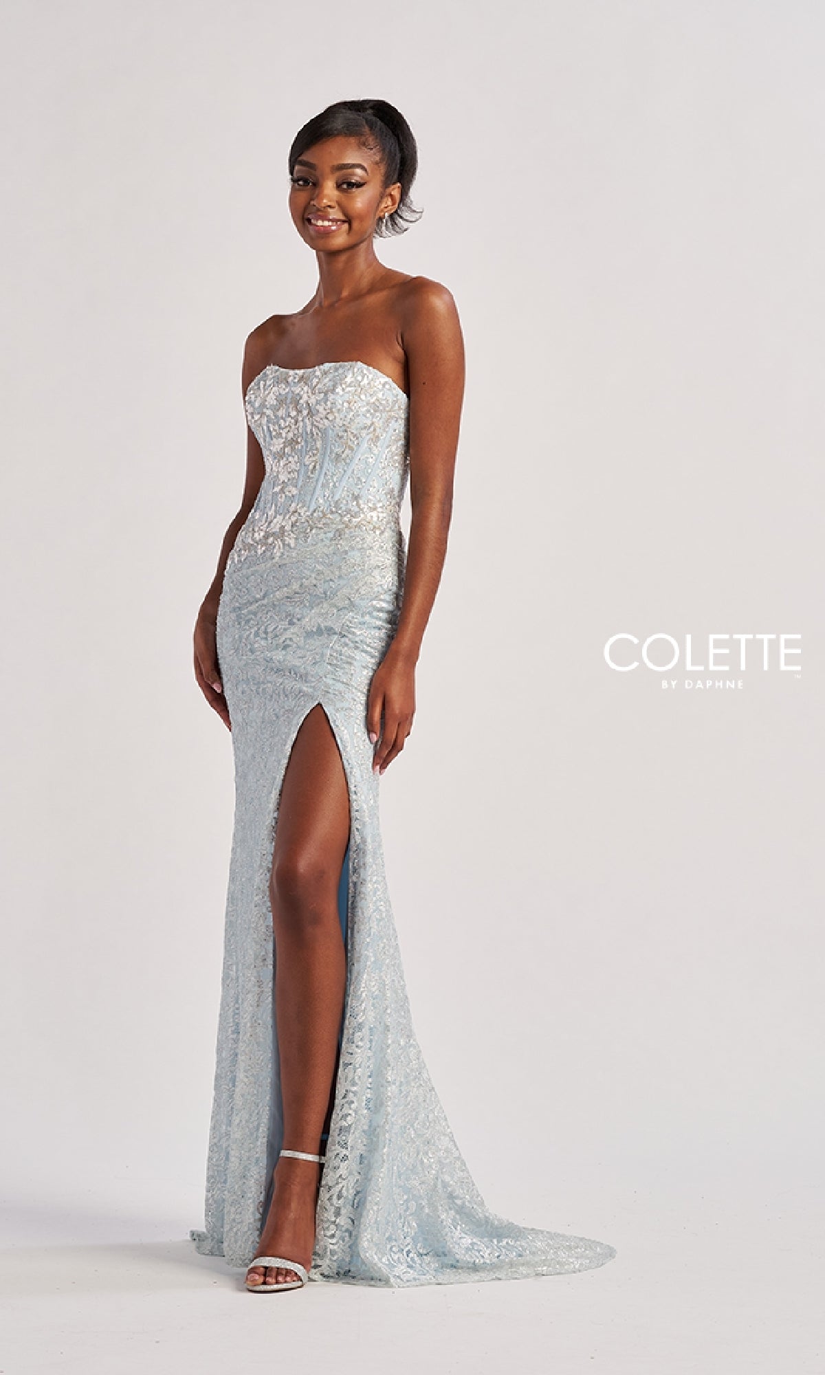 A woman stands smiling against a plain background, wearing the Long Prom Dress: Colette CL8595, a strapless silver-blue lace gown with a high slit.